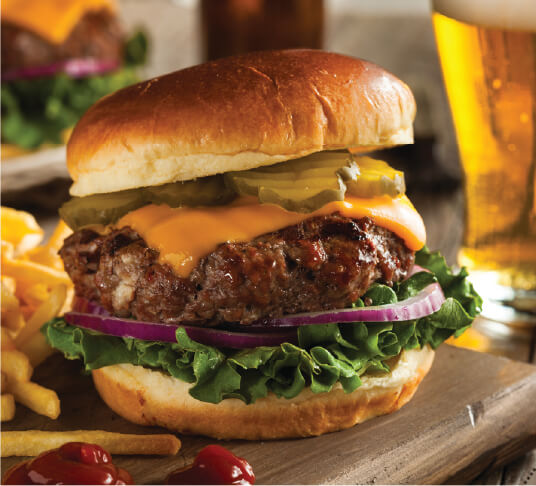 large cheeseburger topped with pickles, red onoin and lettuce on a wooden board with crispy french fries